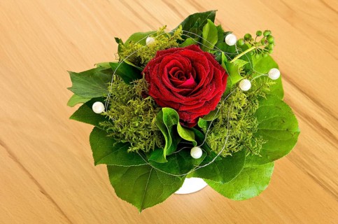Single rose with greens and decorative beads tied into a bouquet, in a vase on a wooden table top