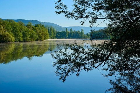 A calm body of water with surrounding trees under a clear sky, the mountains are visible in the