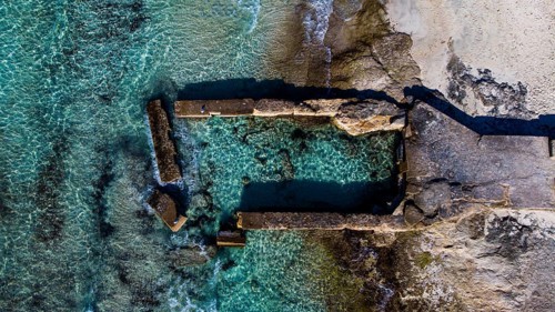 An aerial view of ruins with adjacent rocky coast and clear ocean, natural swimming pool, aerial