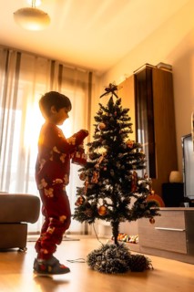 Young boy in pajamas carefully hanging ornaments on a small christmas tree, preparing for joyous