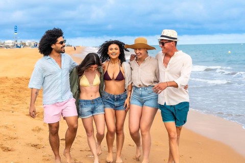 Young multi ethnic friends walking barefoot along a sunny beach, laughing and arm in arm, enjoying