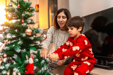 Mother and young son in pajamas decorate a small christmas tree together in a cozy living room,