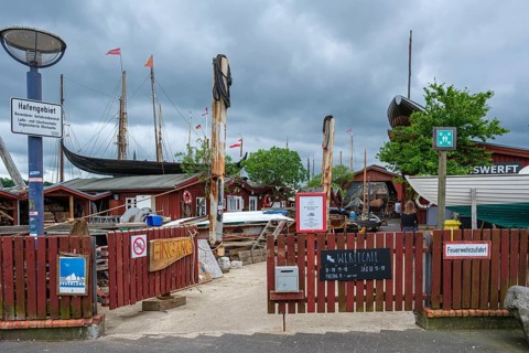 Access to a port area with rocking chairs, wooden buildings and a boat in the background,