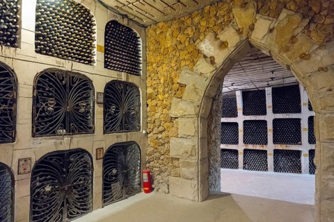A stone arch separates two sections of a wine cellar filled with wine bottles on shelves, wine