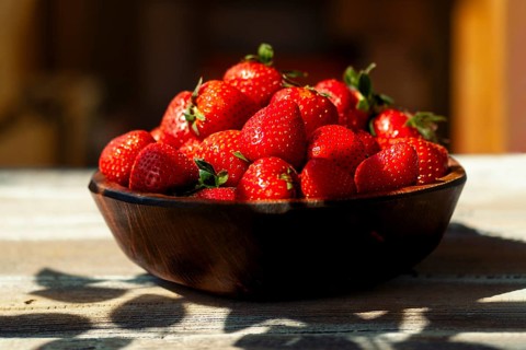 Close-up view of bowl on wooden table with plenty of fresh strawberries inside. Bright daylight