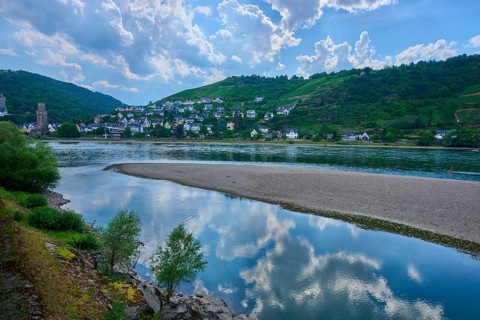 A quiet village stretches across hills on the banks of a river with a sandbar, under a cloudy sky,