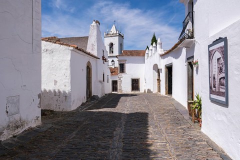 Paved old town street with white buildings and a church, blue sky in the background, Monsaraz,