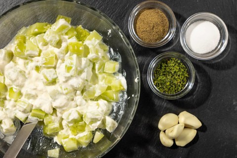 Bowl with sliced cucumber and sour cream, spices and garlic next to it