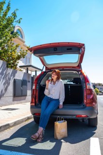 Woman sitting on open suv trunk eating an apple, relaxed after grocery shopping on a sunny day,