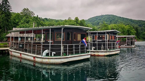 Wooden boats with the Croatian flag are moored on a calm lake against a wooded backdrop, ferry in