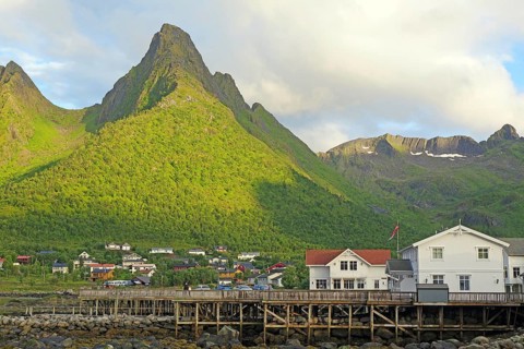 A village with white houses and a wooden bridge lies in front of a majestic mountain with a green