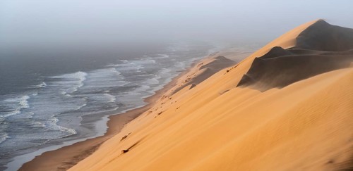 Sand dunes with wave patterns and sea in evening light, wind blowing sand across the dunes,