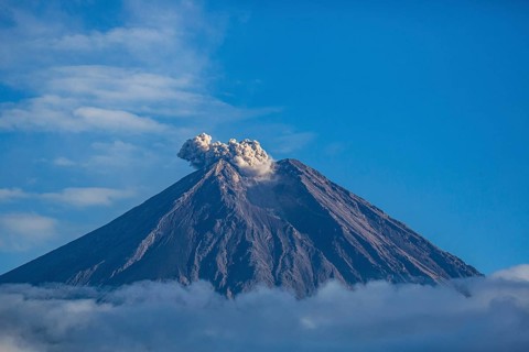 The volcano expels smoke from its summit, under a clear blue sky with surrounding clouds, Smoking