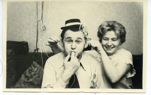 A man wearing a party hat blows impressively streamers while a woman behind him laughs, kaneval,