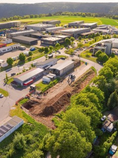 An aerial view of a construction site next to industrial buildings, surrounded by green trees and