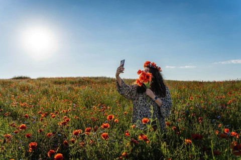 Woman taking selfie in blooming poppy (Papaver rhoeas) field while wearing floral crown and holding