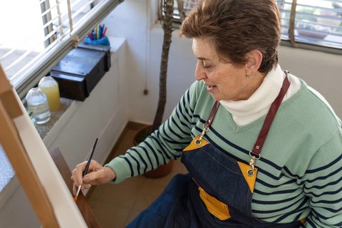 Happy senior woman painting on a canvas at a wooden easel in her bright home art studio. Active