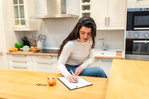 Young woman in a modern kitchen organizing a budget on a clipboard while placing savings in a piggy