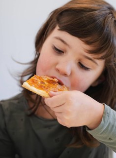 A young child with brown hair takes a bite of a delicious slice of cheese pizza. The scene captures