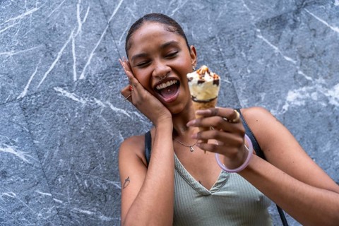 Young woman with dark skin and a nose piercing expressing joy and laughter, holding an ice cream