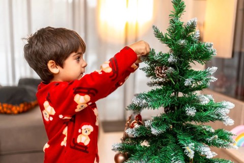Young child in festive pajamas intently placing an ornament on a small christmas tree, celebrating