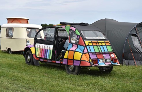 A brightly painted Citroën 2CV stands in front of caravans and tents, colorful view, Citroën 2CV at