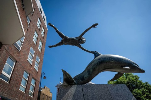 Modern sculpture boy with dolphin by David Wynne against a blue sky, Chelsea Embankment, London,