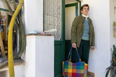 Older woman standing beside the entrance of her home with a colorful shopping bag, representing