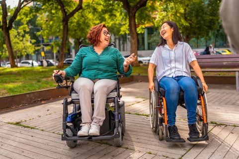 Two multiracial women with disability in wheelchair laughing talking together in a city park