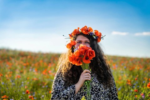 A plus size woman wearing a wreath of wildflowers among a blooming poppy field covers her face with