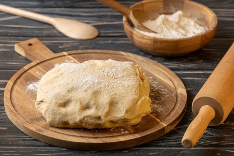 Fresh dough on a wooden board next to a bowl of flour and kitchen utensils