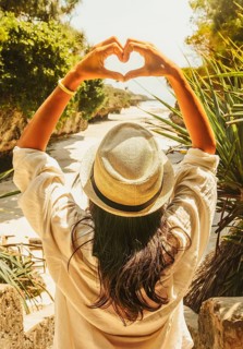 Young Woman tourist in straw hat framing heart sign with hands overhead looking down at tropical