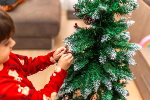 Young boy in red pajamas carefully hangs ornaments on a festive christmas tree at home, smiling