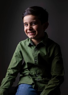 A young boy in a green shirt and jeans sits against a dark background, smiling warmly. The portrait