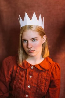 A young teenage girl with blonde hair wears a handmade paper crown and a vintage-style red dress.