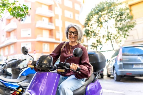 Mature woman with gray hair and glasses smiling at the camera, driving an electric scooter in an