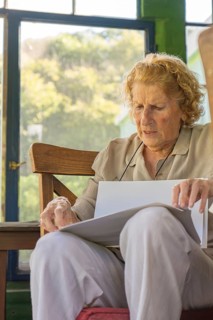 Elderly woman sitting on a wooden chair in a porch, holding and reading documents with a serious