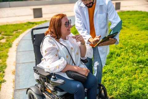 Doctor showing medical information to a smiling woman sitting in a wheelchair in a park, promoting