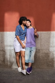 A loving multiethnic LGBTQ+ couple stands closely together, embracing and smiling against a warm
