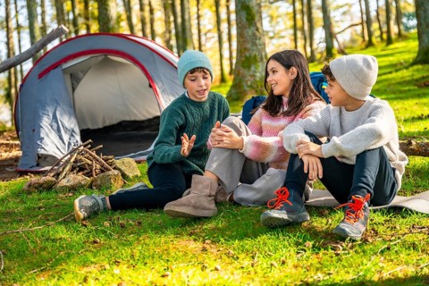Two caucasian brothers and sister talking sitting next to camping tent in the forest