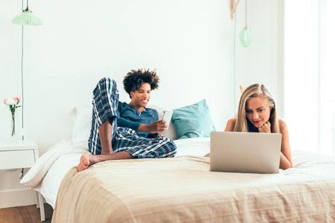 Two friends enjoy a relaxed morning in a bright bedroom. One uses a laptop while the other is on a