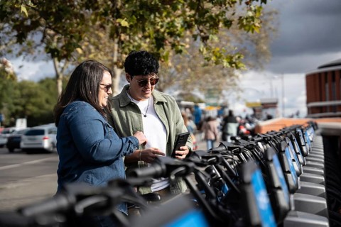 A joyful lesbian couple explores a row of rental bikes in a vibrant city setting. Background