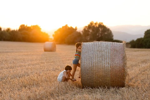 Two young children, a boy and a girl, are joyfully playing beside a large hay bale in a vast golden