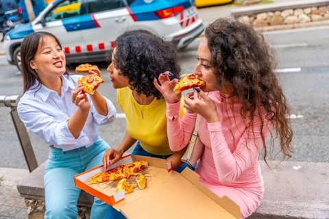 Three multiracial women sharing pizza sitting together in the street