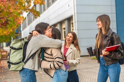 Four cheerful young women students hug and laugh on a university campus amid autumn leaves,