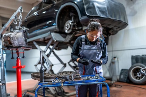 Female mechanic wearing gloves and safety glasses examining car parts with a car on a hydraulic