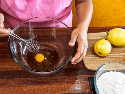 A cook prepares lemon cookie dough by beating an egg in a glass bowl. Nearby, fresh lemons and