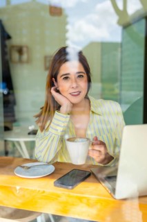 Young woman behind a cafe window, smiling as she works on her laptop with a warm cup of coffee,