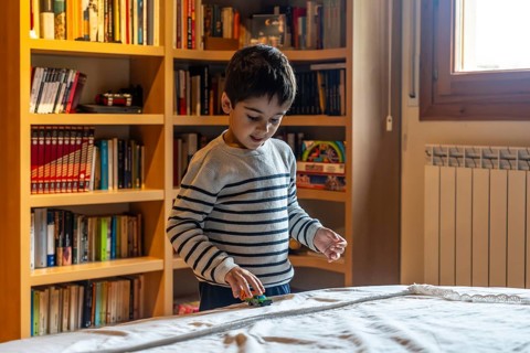 Young boy happily playing with a toy car on a white duvet cover, standing in front of a wooden