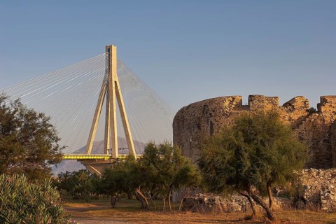 Ruins of an old city near a modern bridge, surrounded by trees in warm evening light, Rio-Antirrio